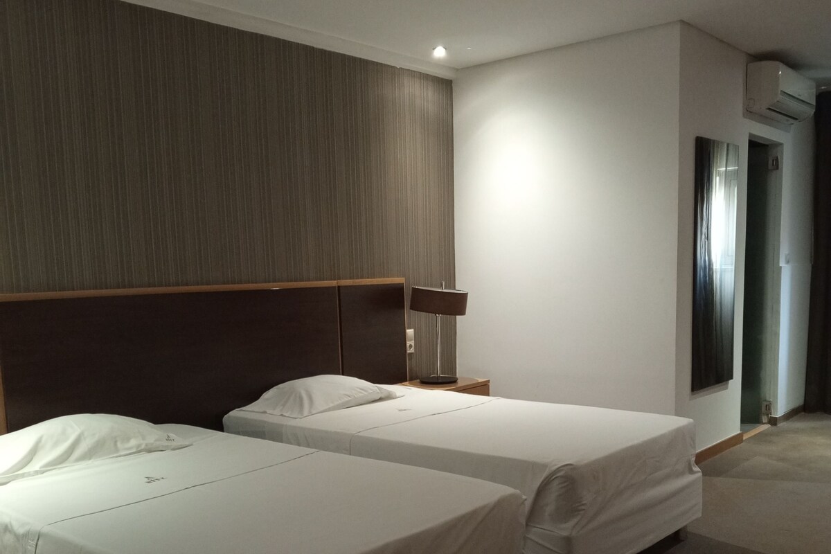 Ein Hotelzimmer mit zwei Betten, einem Kopfteil, einem Nachttisch, einer Lampe und einer Klimaanlage.