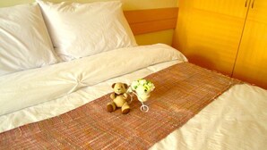 A bed with white linens, a brown blanket, a teddy bear, and a small vase with flowers. - Dariva Place (Pattaya)