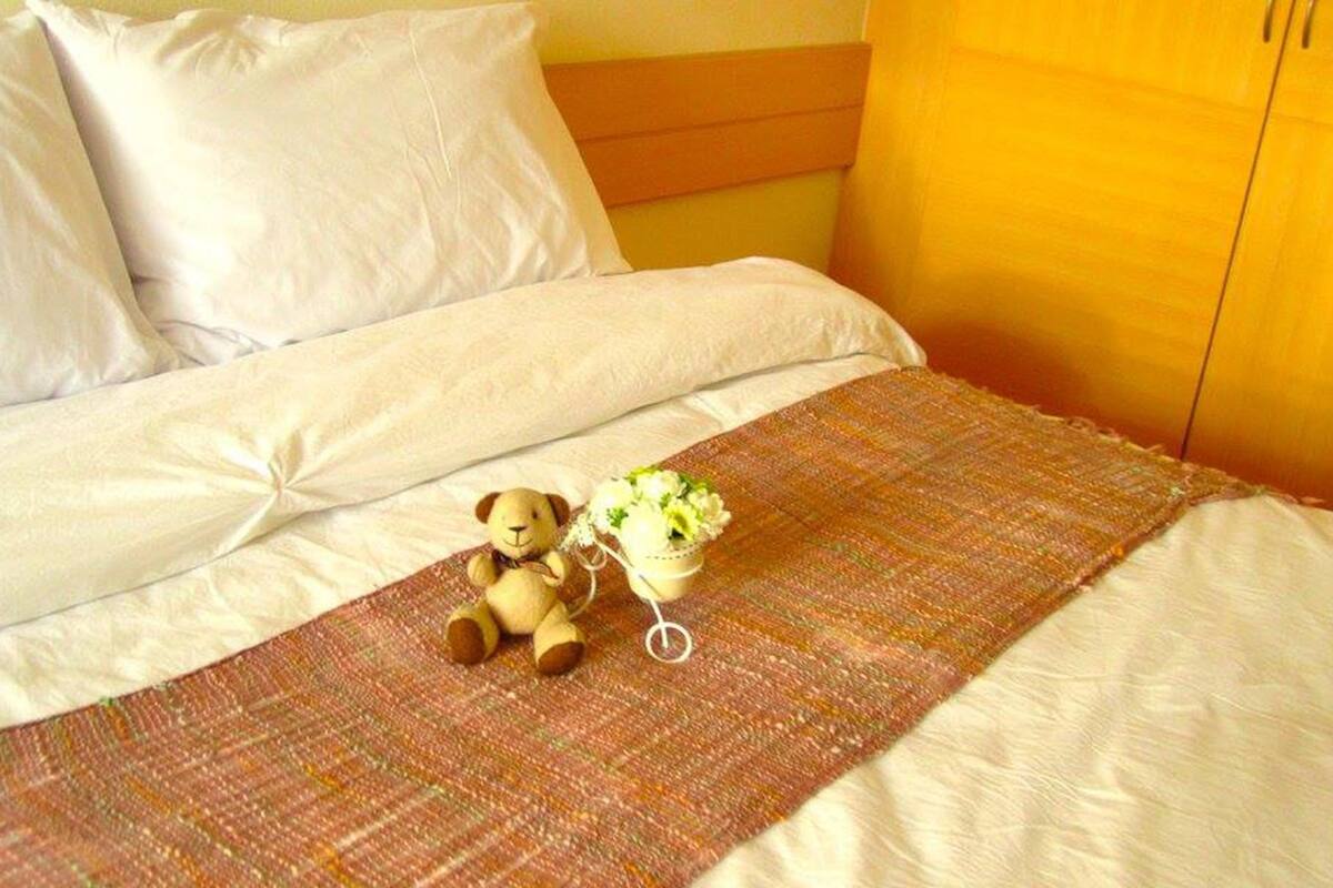 Un lit avec du linge de lit blanc, une couverture marron, un ours en peluche et un petit vase avec des fleurs.