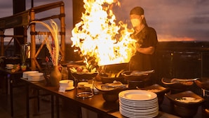 Un cocinero preparando comida con un fuego grande en una cocina rústica al aire libre.