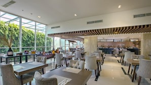 Un restaurante espacioso con techos altos, ventanas amplias y una zona de cocina central. Las mesas están dispuestas para comer, y el espacio está bien iluminado con luz natural y artificial.