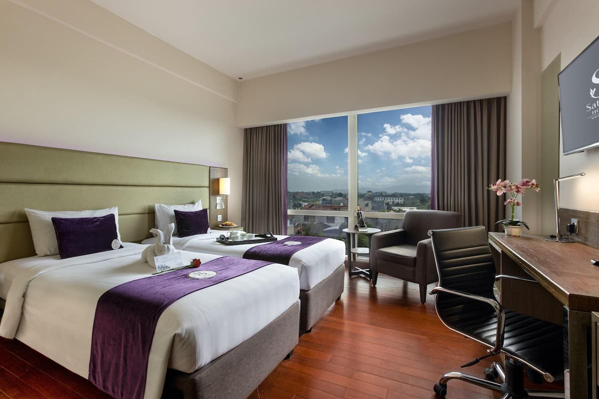 Habitación de hotel con dos camas, un escritorio, una silla, una mesita y vistas a la ciudad.