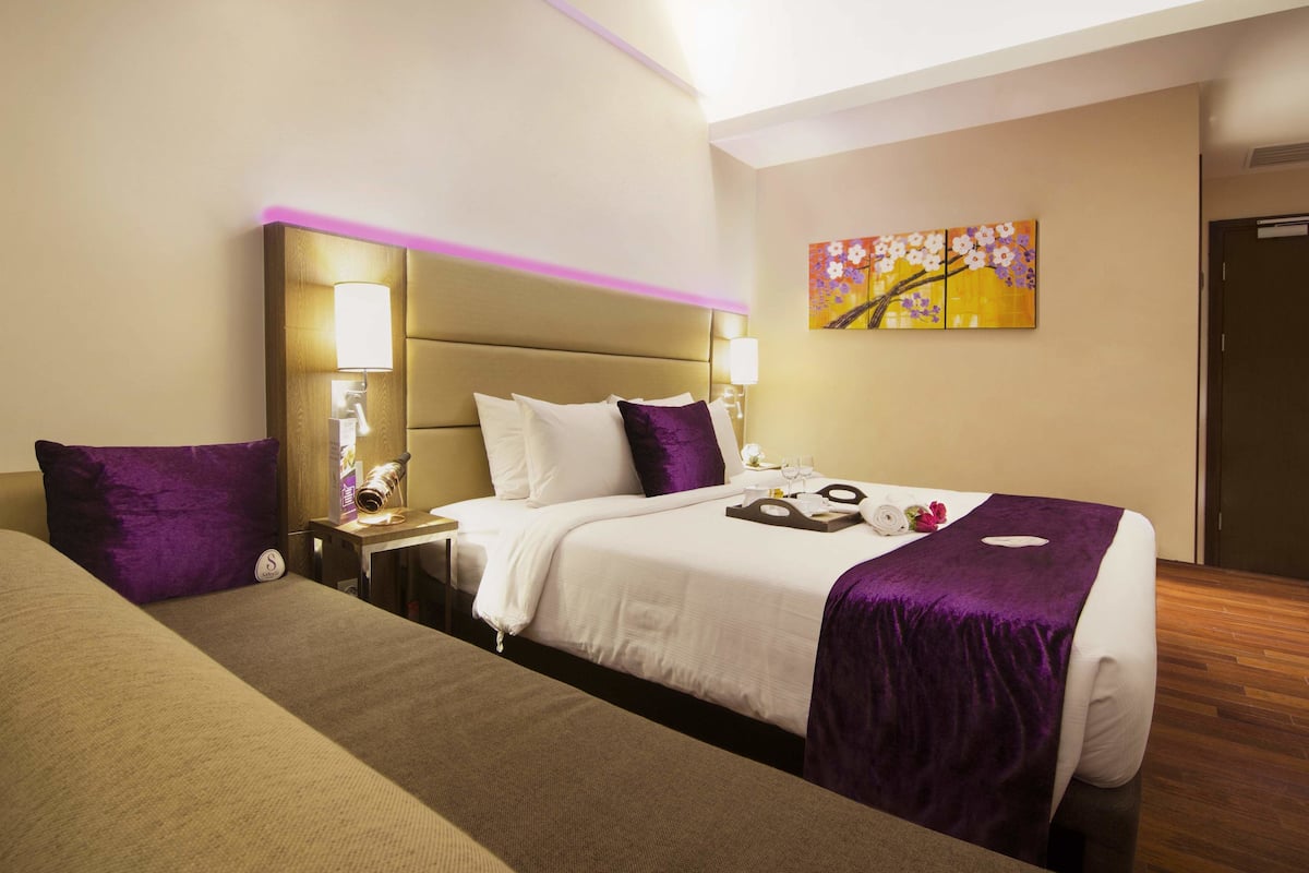 Una habitación de hotel con una cama grande, una mesita de noche con lámpara, una mesita auxiliar y un cuadro en la pared.