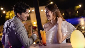 Una pareja sentada a una mesa, con una bebida frente a ellos, bajo luces de cuerda.