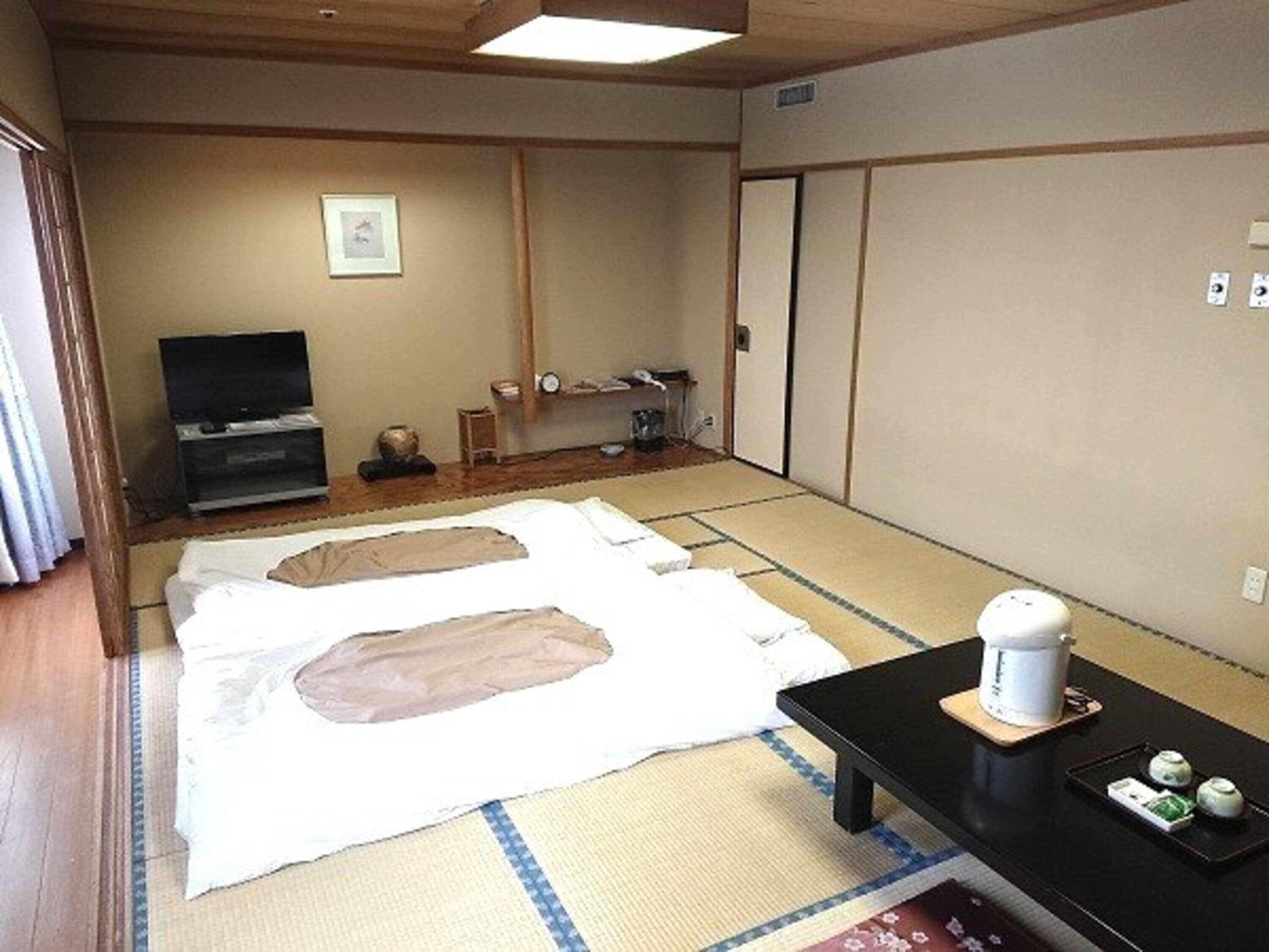 A traditional Japanese room with tatami mats, a futon bed, a television, and a small table with a kettle and cups.