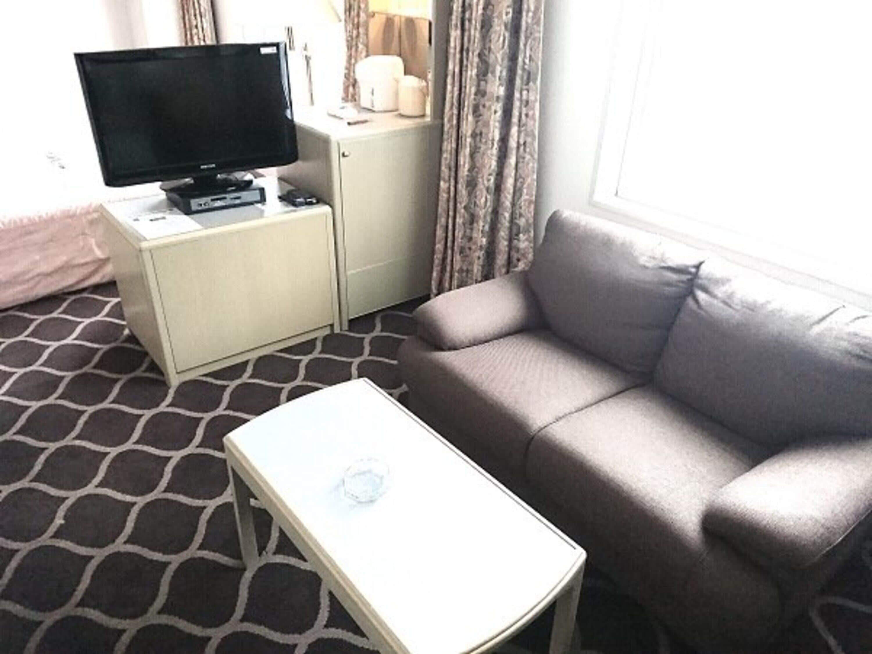 A hotel room with a grey sofa, a small white table, a TV on a cabinet, and patterned carpet.