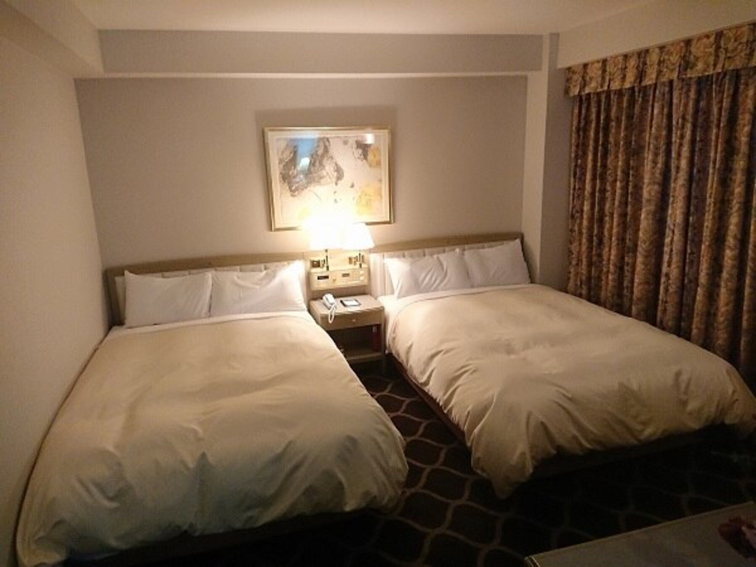 A hotel room with two single beds, a nightstand with a lamp, and a painting on the wall.