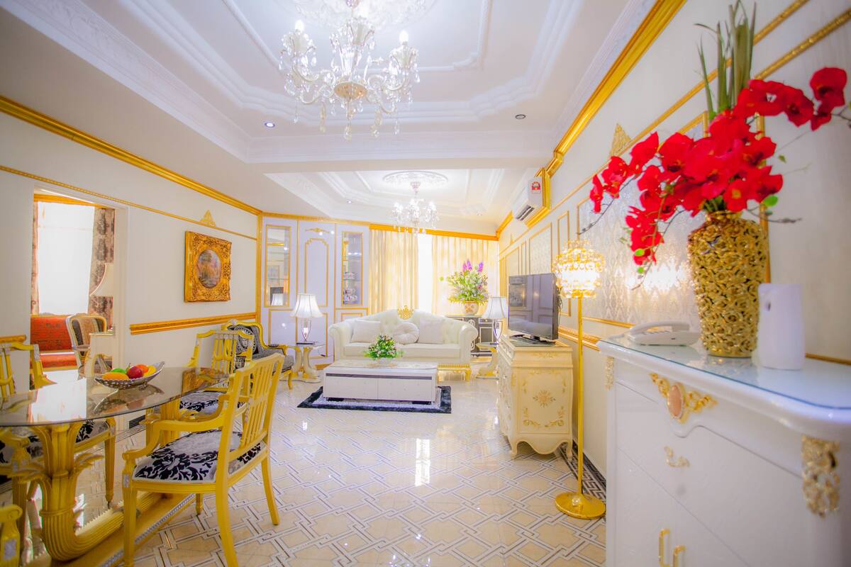 A luxurious living room with a chandelier, a white sofa, a dining table with yellow chairs, and a decorative vase with red flowers.