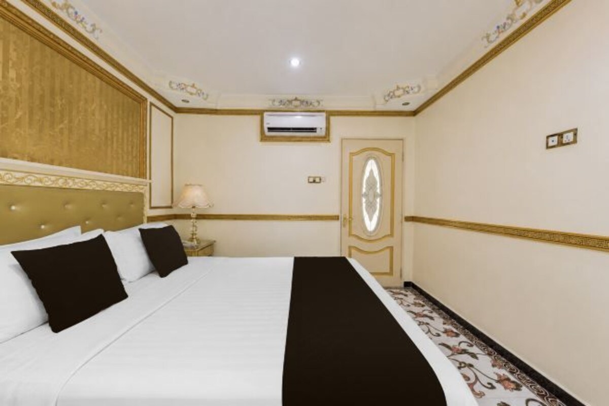 A hotel room with a large bed, a bedside table with a lamp, an air conditioning unit, and a door with a mirror.