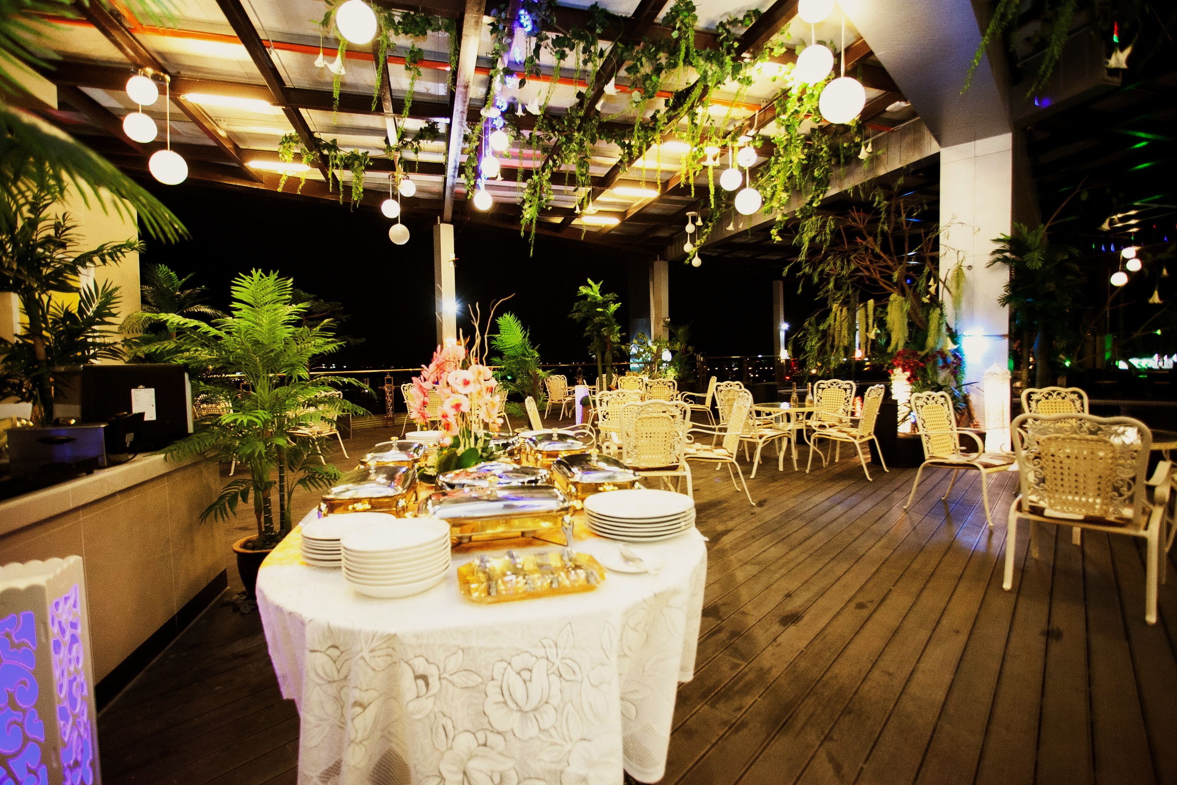An outdoor dining area with round tables set for a meal, surrounded by wicker chairs, and decorated with hanging plants and lights.