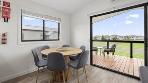 A modern room with a round wooden table, grey chairs, a fire extinguisher, and a large window overlooking a deck with outdoor furniture. - Swan Bay Holiday Park (Swan Bay)