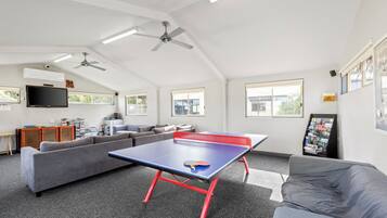 En rummelig opholdsstue med et pingpongbord, grå sofaer, et fladskærms-TV og en loftsvifte.