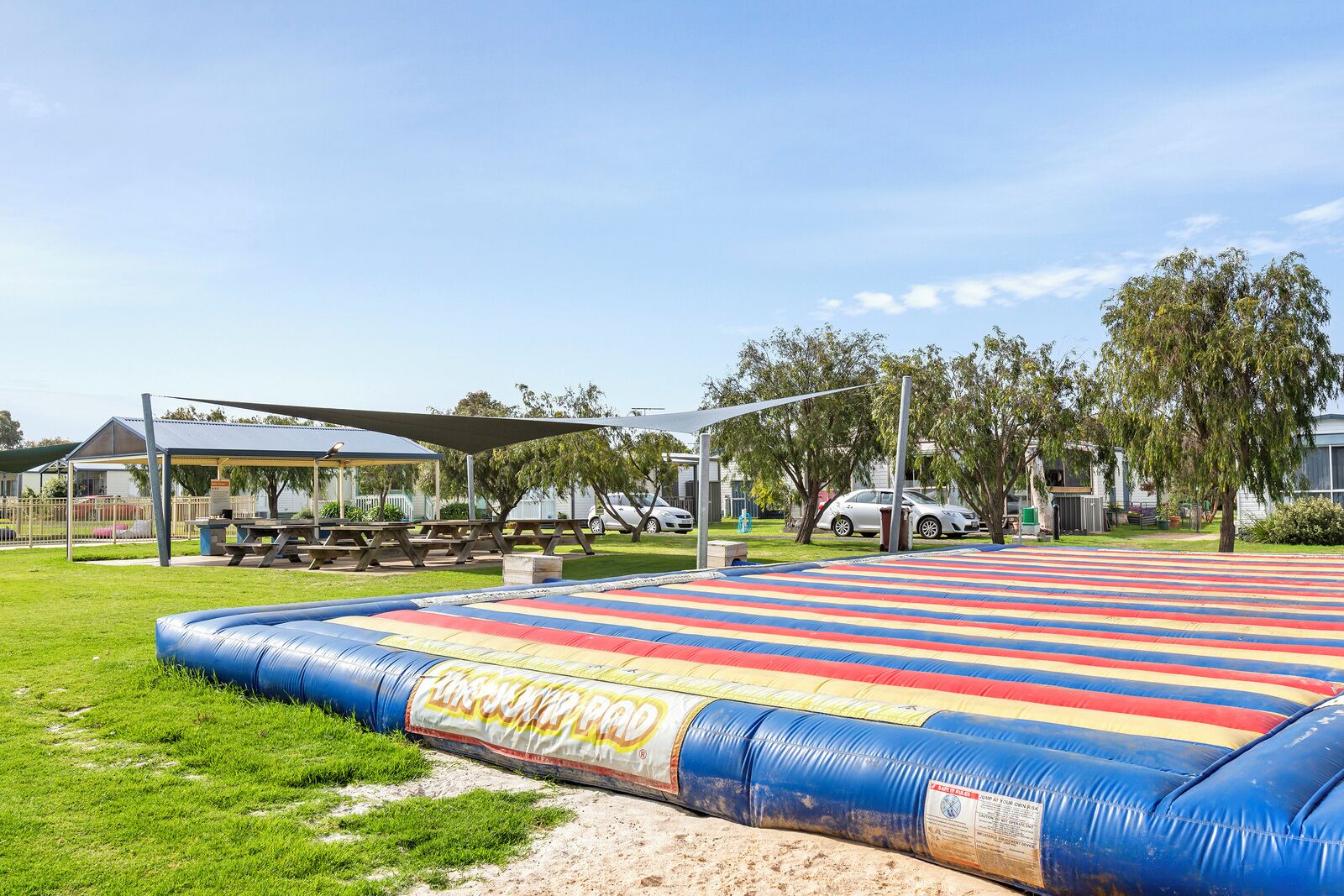 Un espace verdoyant comprenant un grand tapis de saut gonflable, des tables de pique-nique et un pavillon abrité.