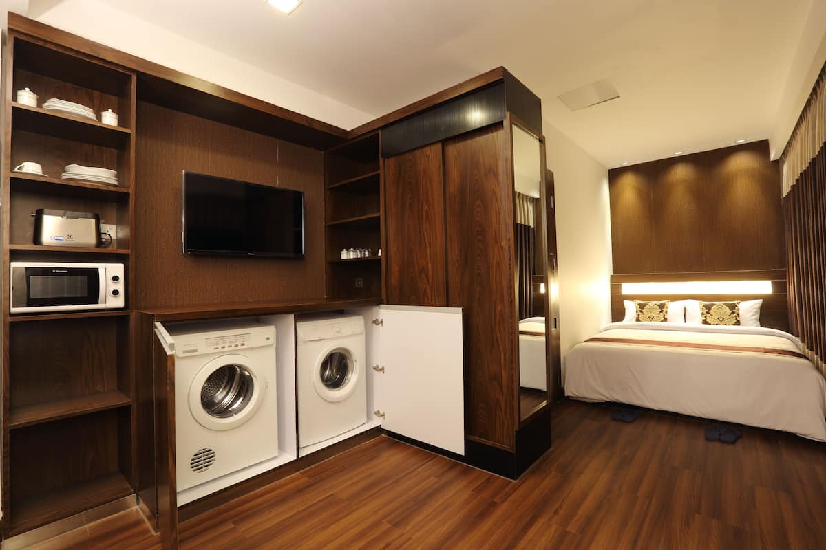 A compact hotel room with a washing machine, microwave, and a bed with a headboard mirror.