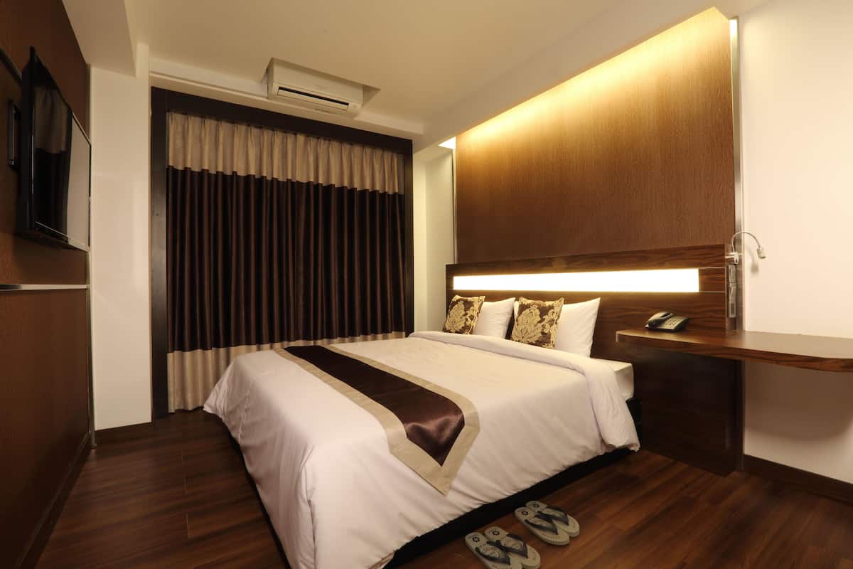 A hotel room with a large bed, a wooden headboard, a bedside table with a phone, and a pair of slippers on the floor.