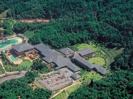 Vista aérea de um resort com vários edifícios, uma piscina e um estacionamento rodeado por uma floresta densa.