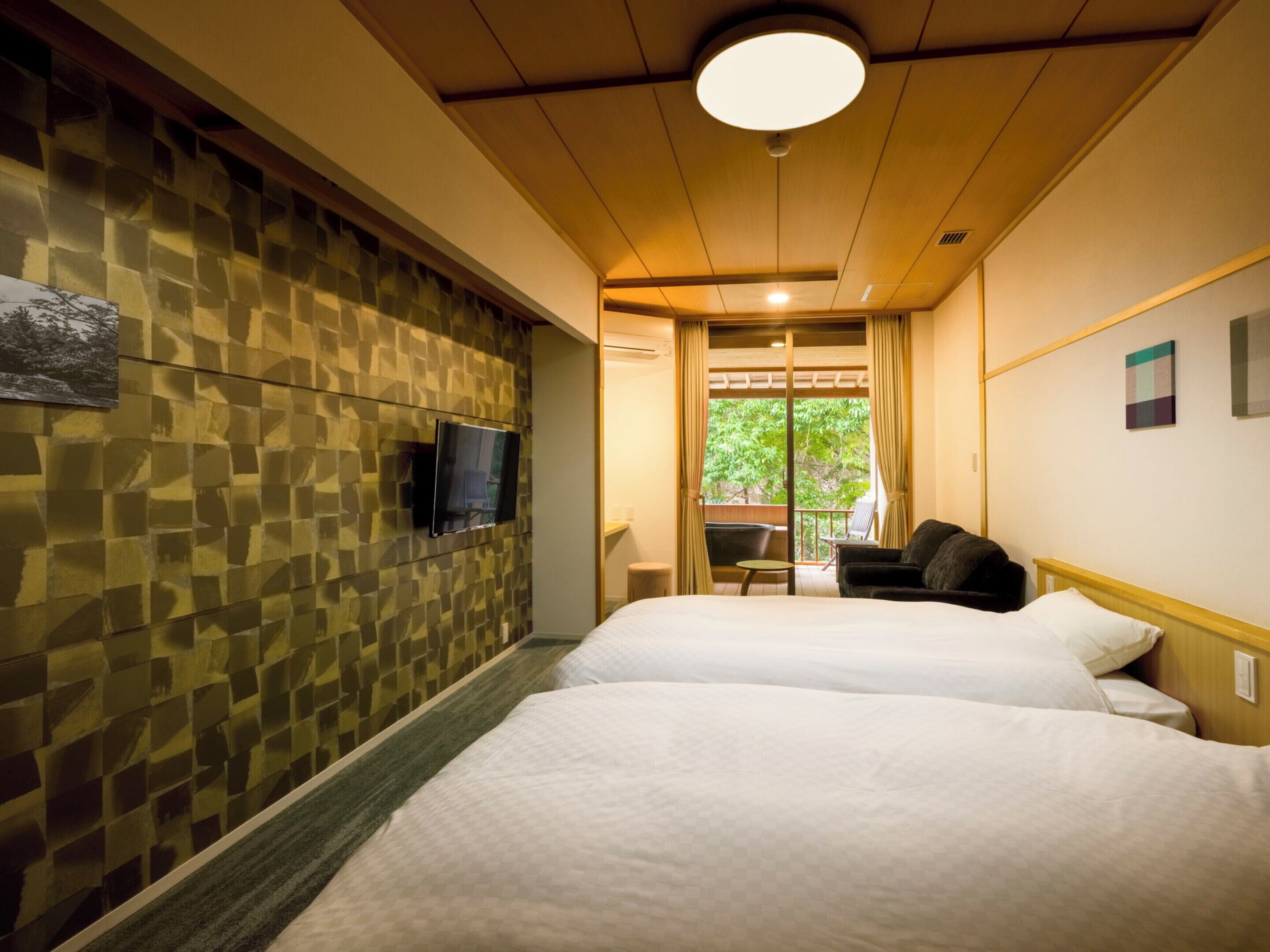 A hotel room with two beds, a television on the wall, and a balcony with a view of trees.