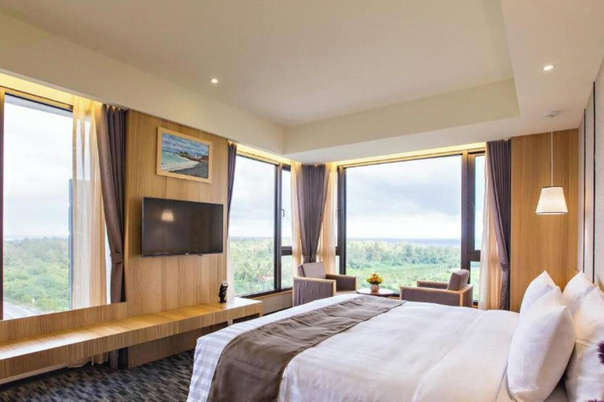 Habitació d'hotel amb un llit gran, televisió, zona de seients i finestrals que ofereixen vista a l'exterior.