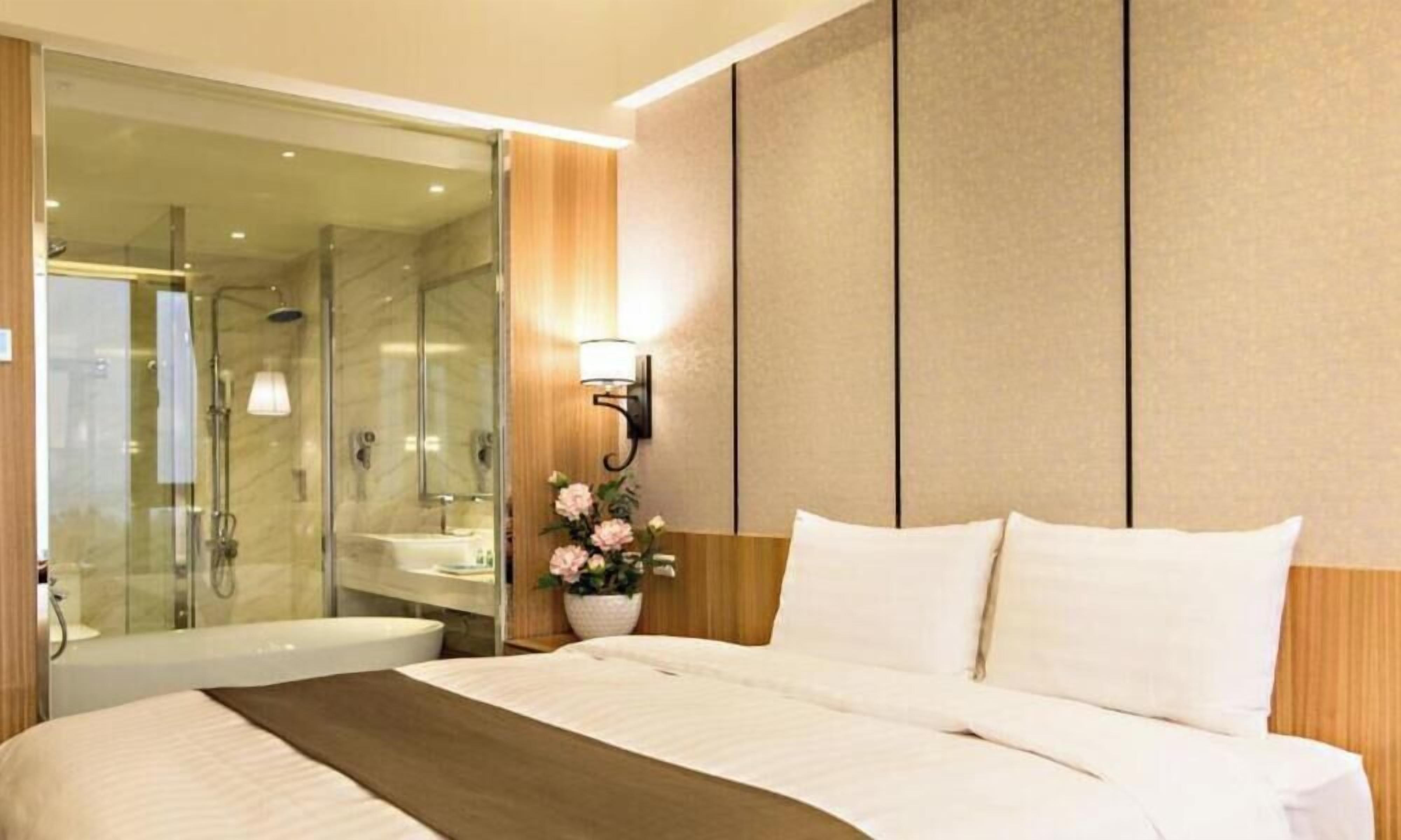 A hotel room with a bed, a bathroom with a shower, and a wall-mounted light fixture.