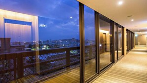 Moderni hotellikäytävä, jossa on suuret ikkunat, puulattia ja näkymä kaupunkiin hämärän aikaan.