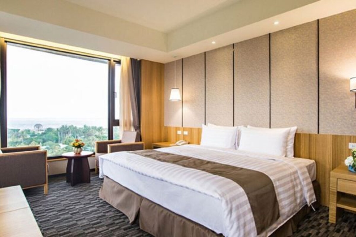 A hotel room with a large bed, a desk, a chair, a nightstand, a lamp, and a view of the city.