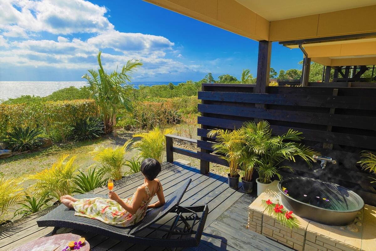 A person relaxing on a lounge chair on a wooden deck with a view of the ocean.