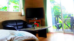 Een kamer met een houten plafond, een flatscreen televisie op een standaard, een bank, een tafel met een bloevas en een balkon met uitzicht op groen.