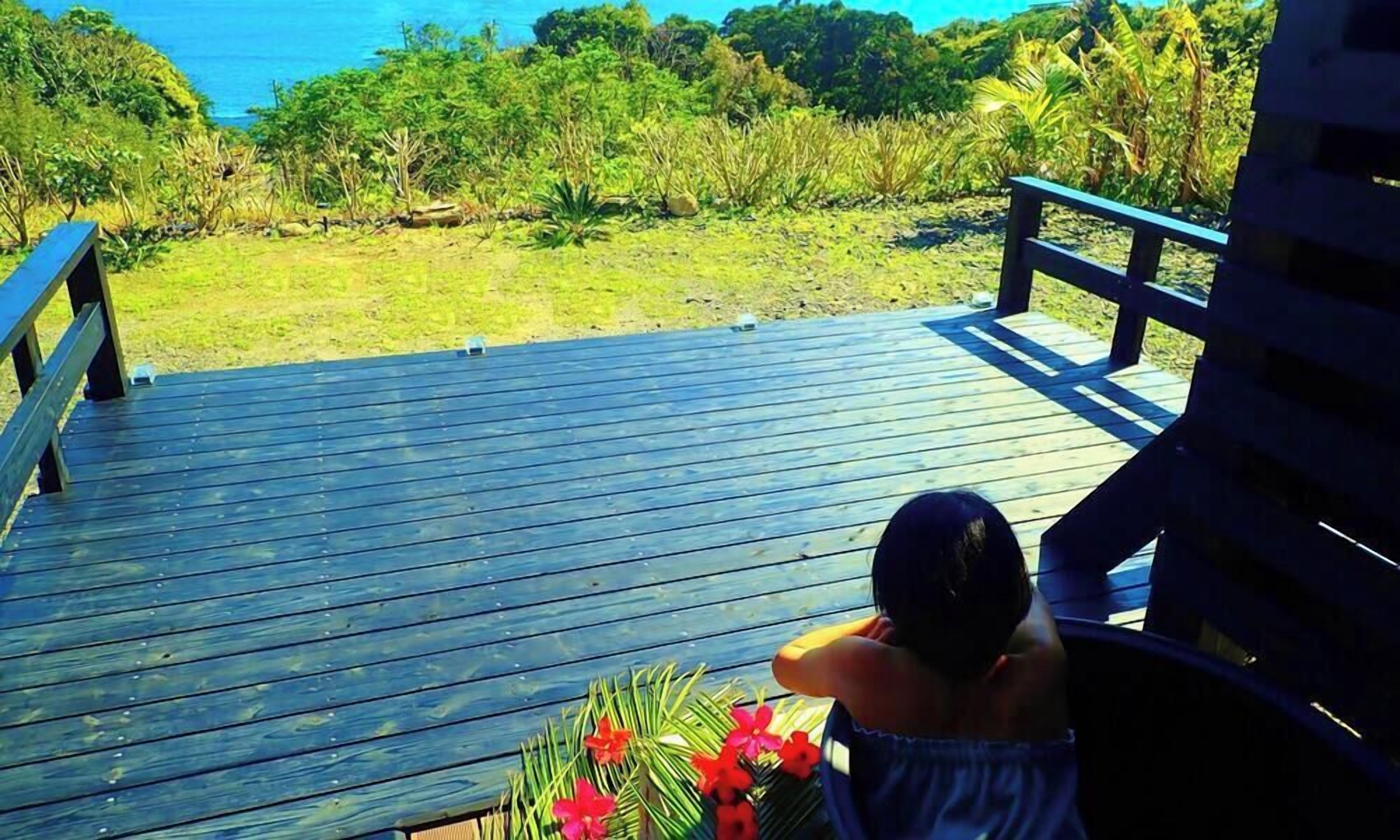 Una terraza de madera con barandilla, con vista a un área verde con vegetación densa y el océano.