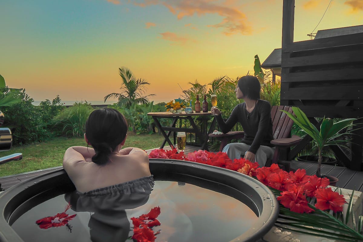 A hot tub with red flowers, a table with bottles, and a view of the sunset.