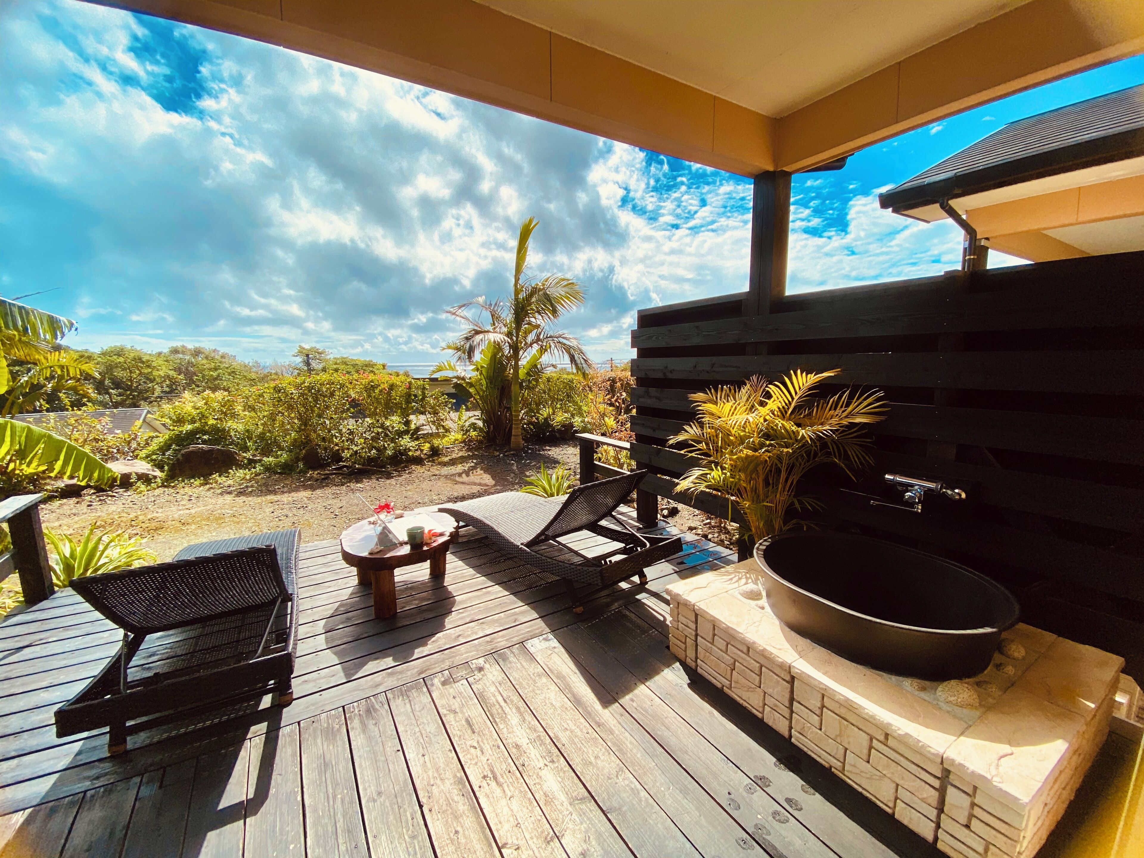 A wooden deck with lounge chairs and a barbecue grill, overlooking a garden with tropical plants and a clear sky.