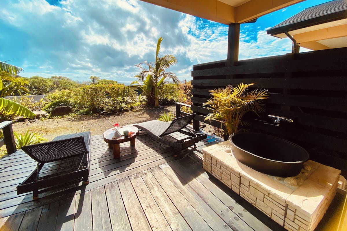 A wooden deck with lounge chairs and a barbecue grill, overlooking a garden with tropical plants and a clear sky.
