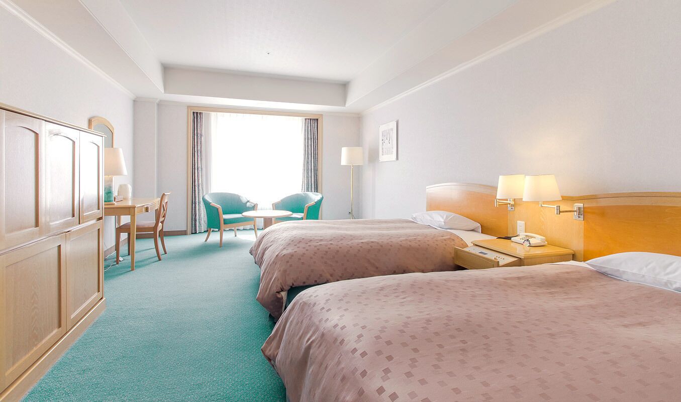 A hotel room with two beds, a desk with a chair, a small table, and a window with curtains.