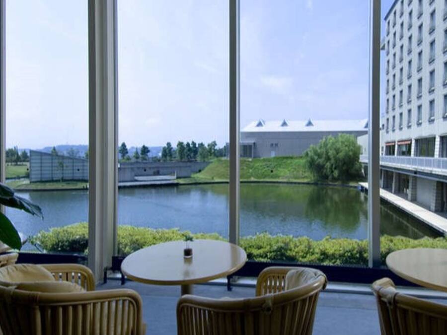 A view from a hotel room with large windows overlooking a body of water, a bridge, and a building across the water. There are wicker chairs and a small table in the foreground.