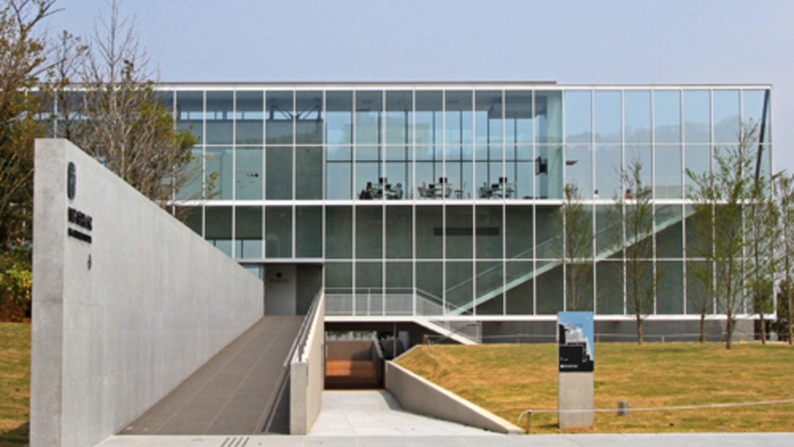 Un edificio moderno con fachada de vidrio, una rampa de hormigón y un área verde al frente.