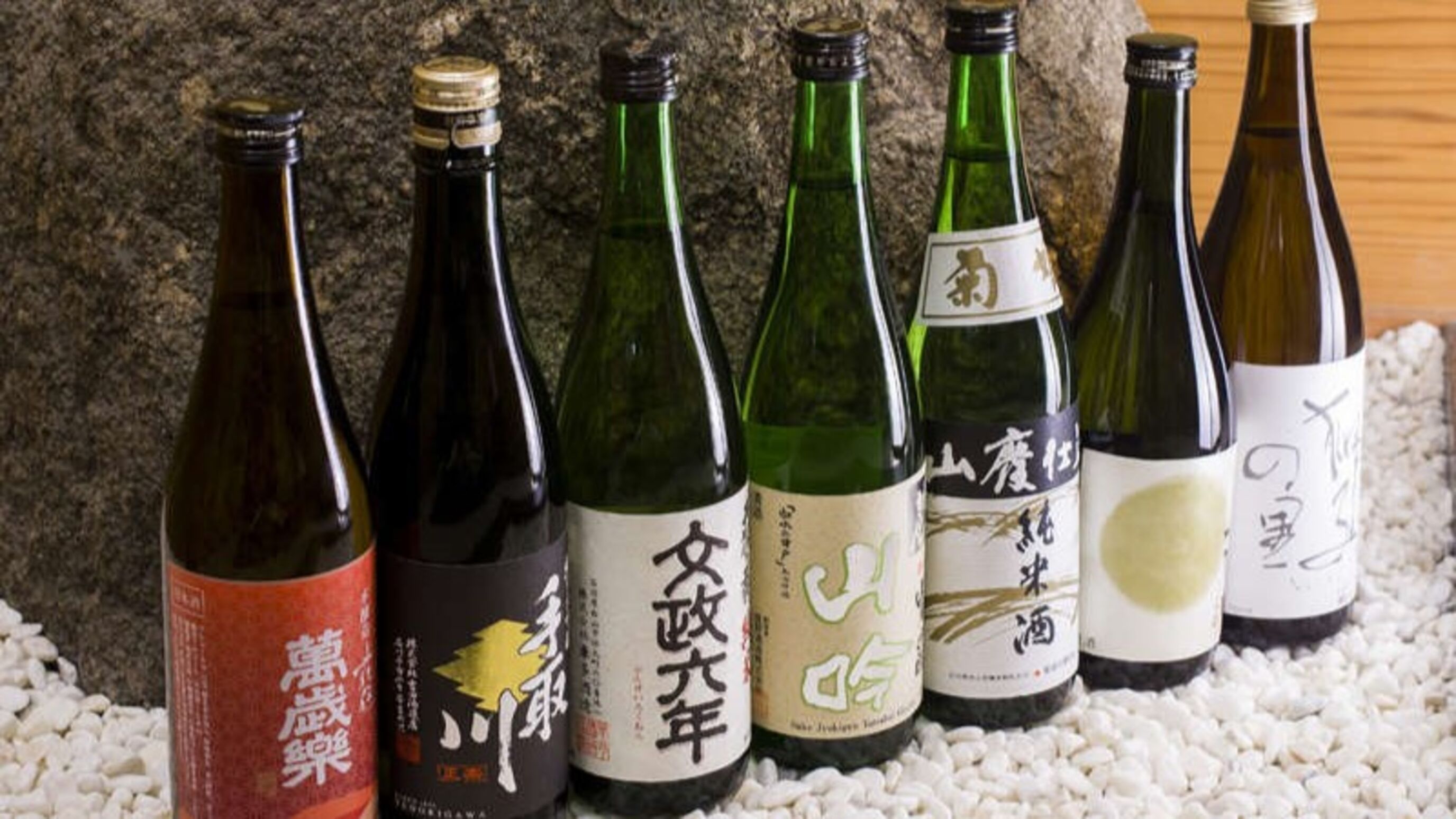A row of six bottles with labels, placed on a bed of white pebbles against a textured background.
