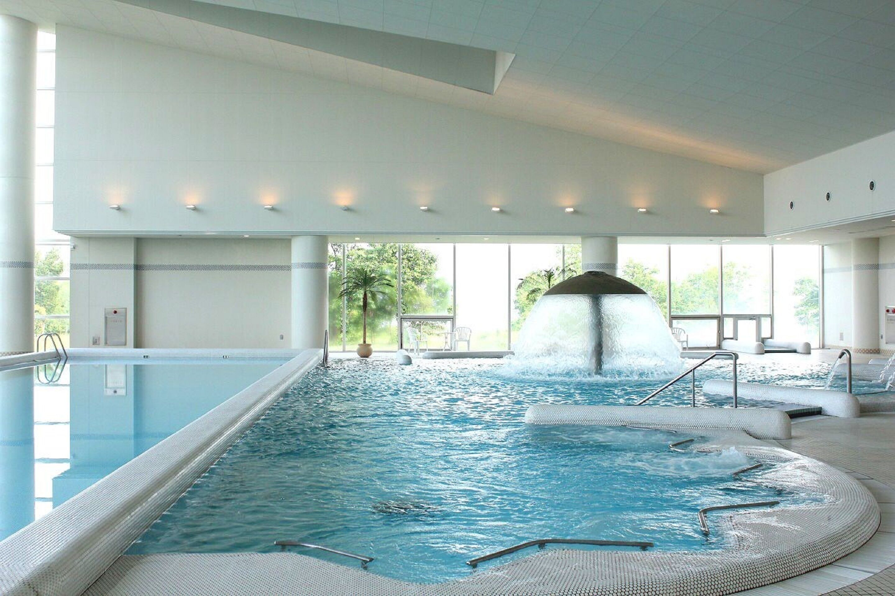 An indoor swimming pool with a waterfall feature, clear glass walls, and a view of greenery outside.