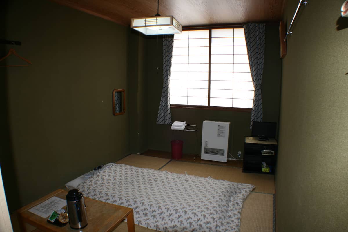 Une petite pièce, sobrement meublée, avec un sol recouvert de tatamis, un futon servant de lit, un radiateur, un téléviseur et une table en bois sur laquelle se trouvent une tasse et un menu.