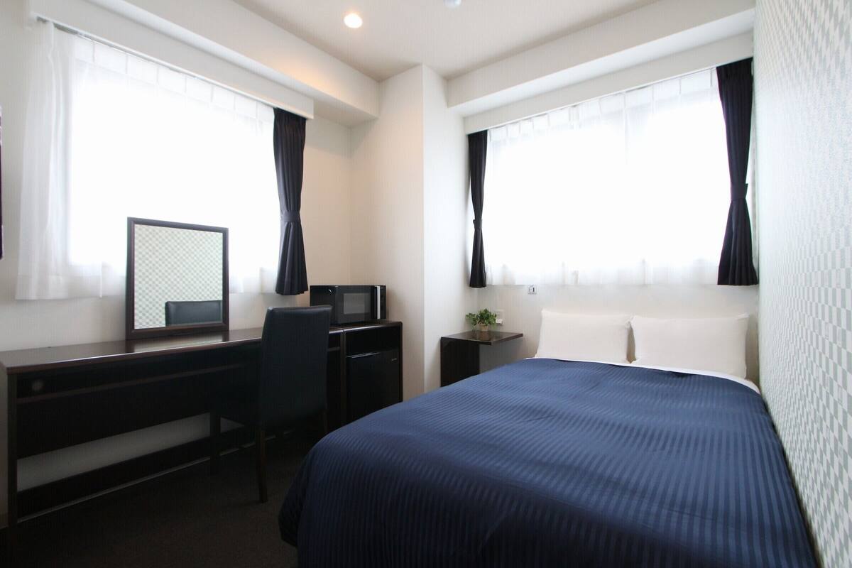 Een hotelkamer met een bed, een bureau met een computer, een stoel, een tafeltje met een plant en twee ramen met gordijnen.
