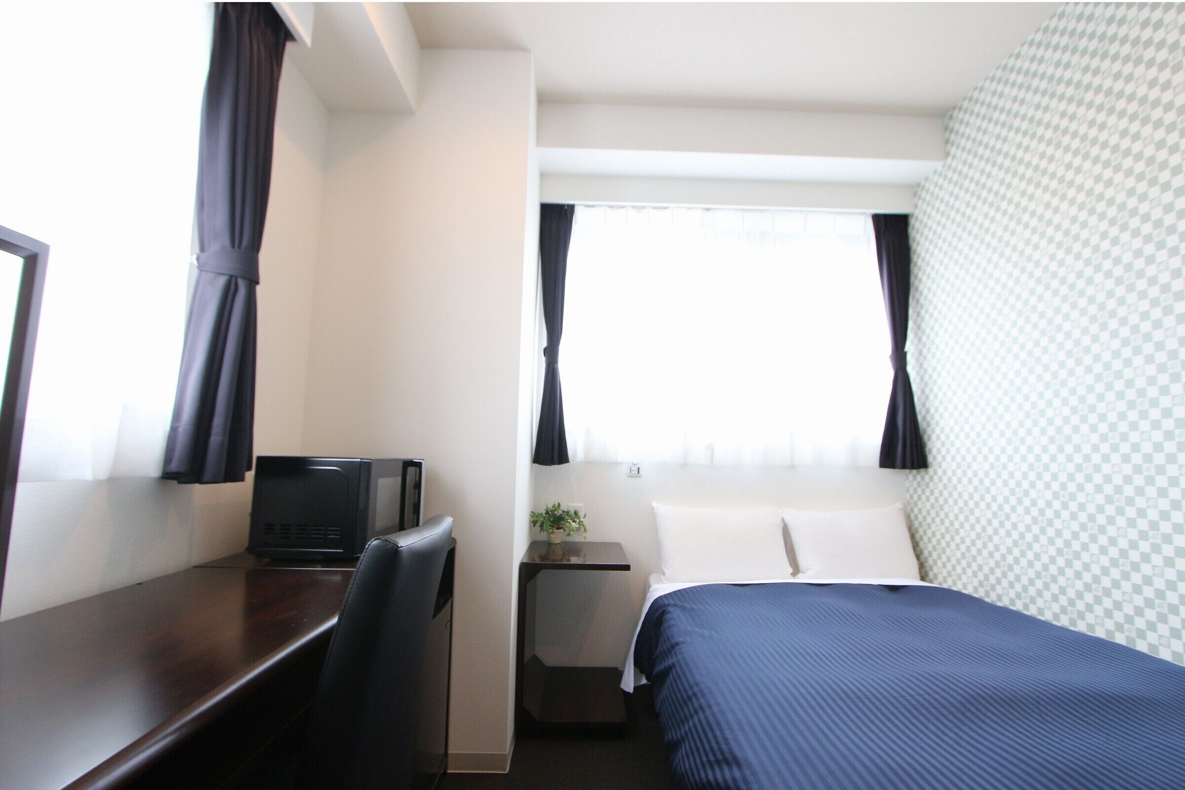 Une chambre d’hôtel avec un lit, un bureau, une chaise, une petite table avec une plante et une fenêtre avec des rideaux.