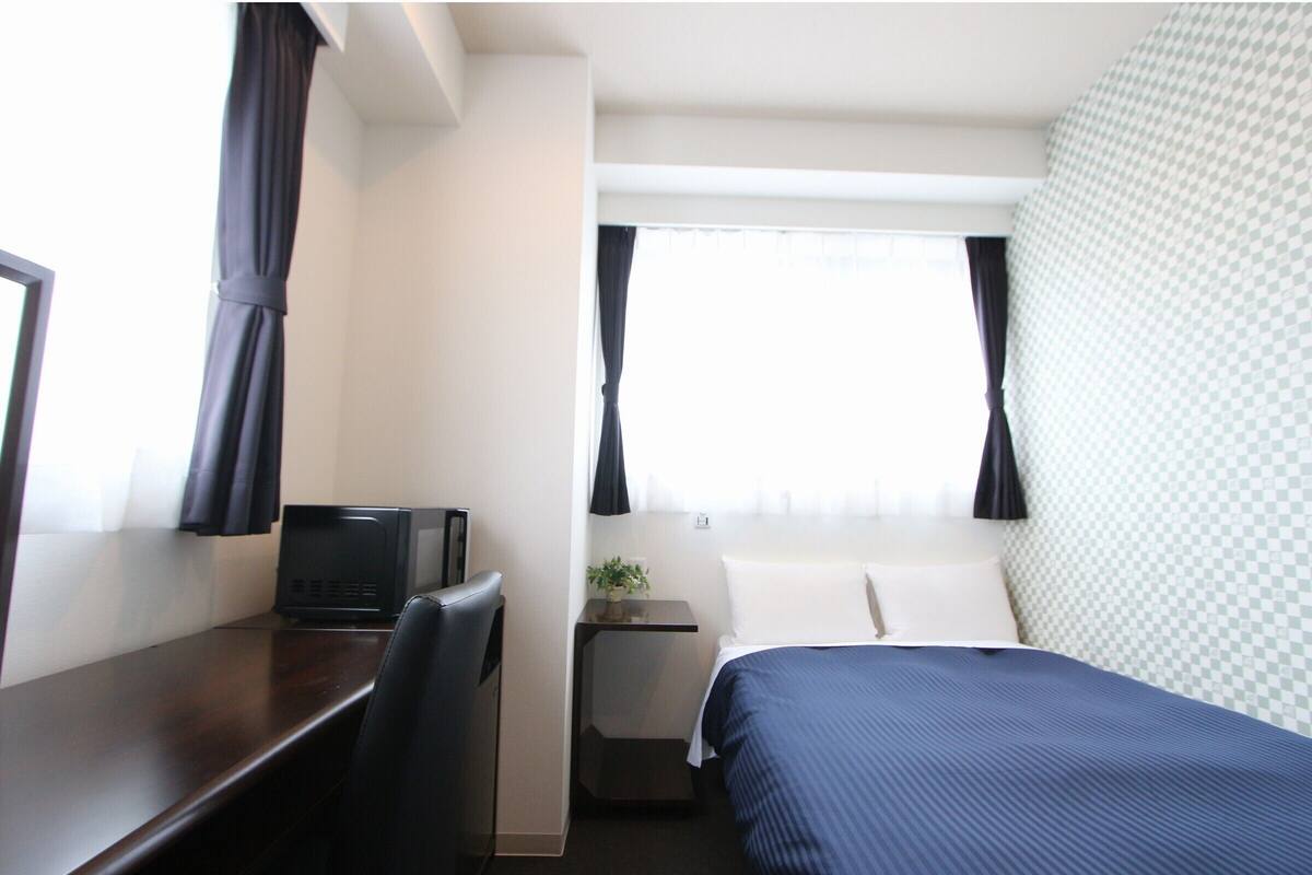 Een hotelkamer met een bed, een bureau, een stoel, een kleine tafel met een plant en een raam met gordijnen.