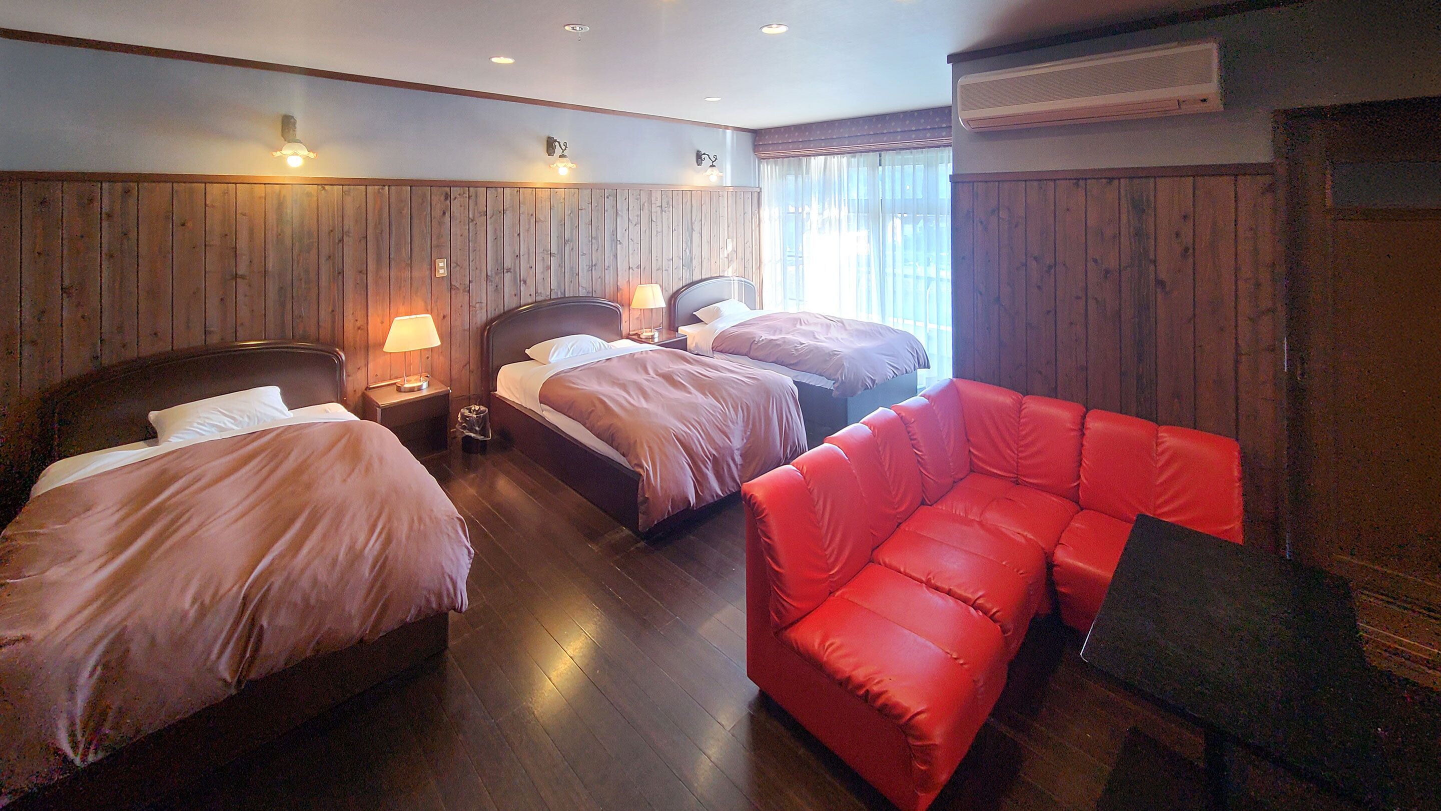 Ein Hotelzimmer mit zwei Betten, einem roten Sofa, Holzvertäfelung und einem Fenster mit Vorhängen.