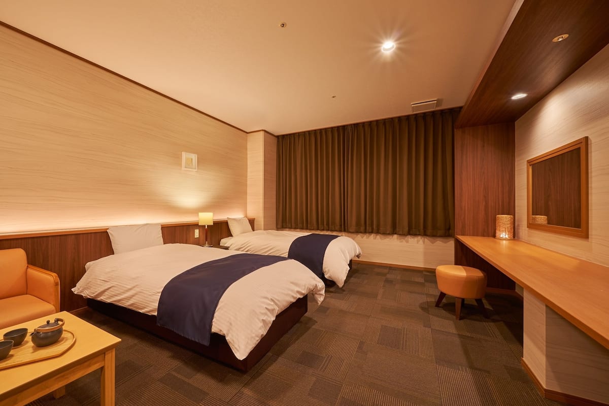 A hotel room with two beds, a sofa, a small table with tea set, and a desk with a lamp.