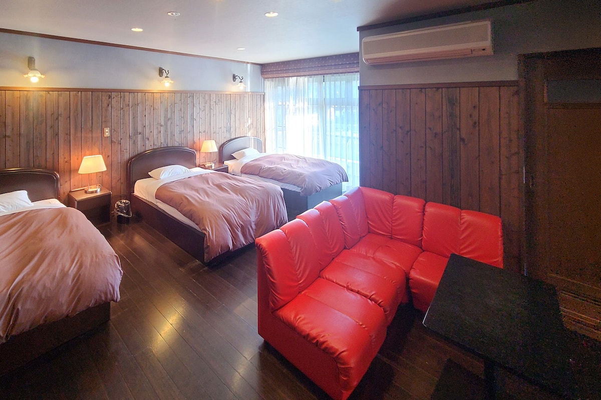 A hotel room with two beds, a red sofa, wooden paneling, and a window with curtains.