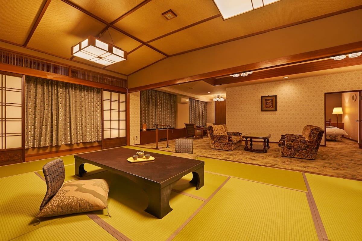 A traditional Japanese room with tatami flooring, a low table with cups, chairs, and a bed in the background.