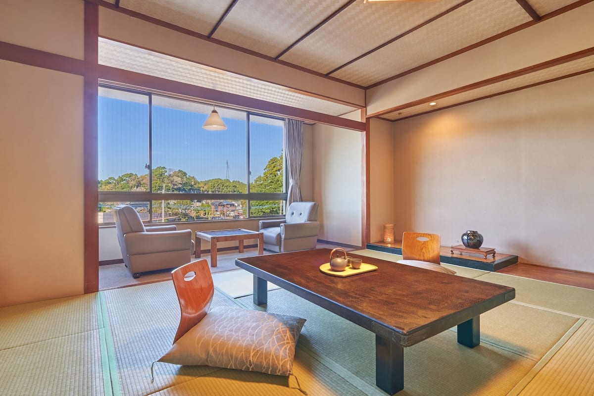 A traditional Japanese room with tatami flooring, a low table with a tea set, and a view of a cityscape through large windows.