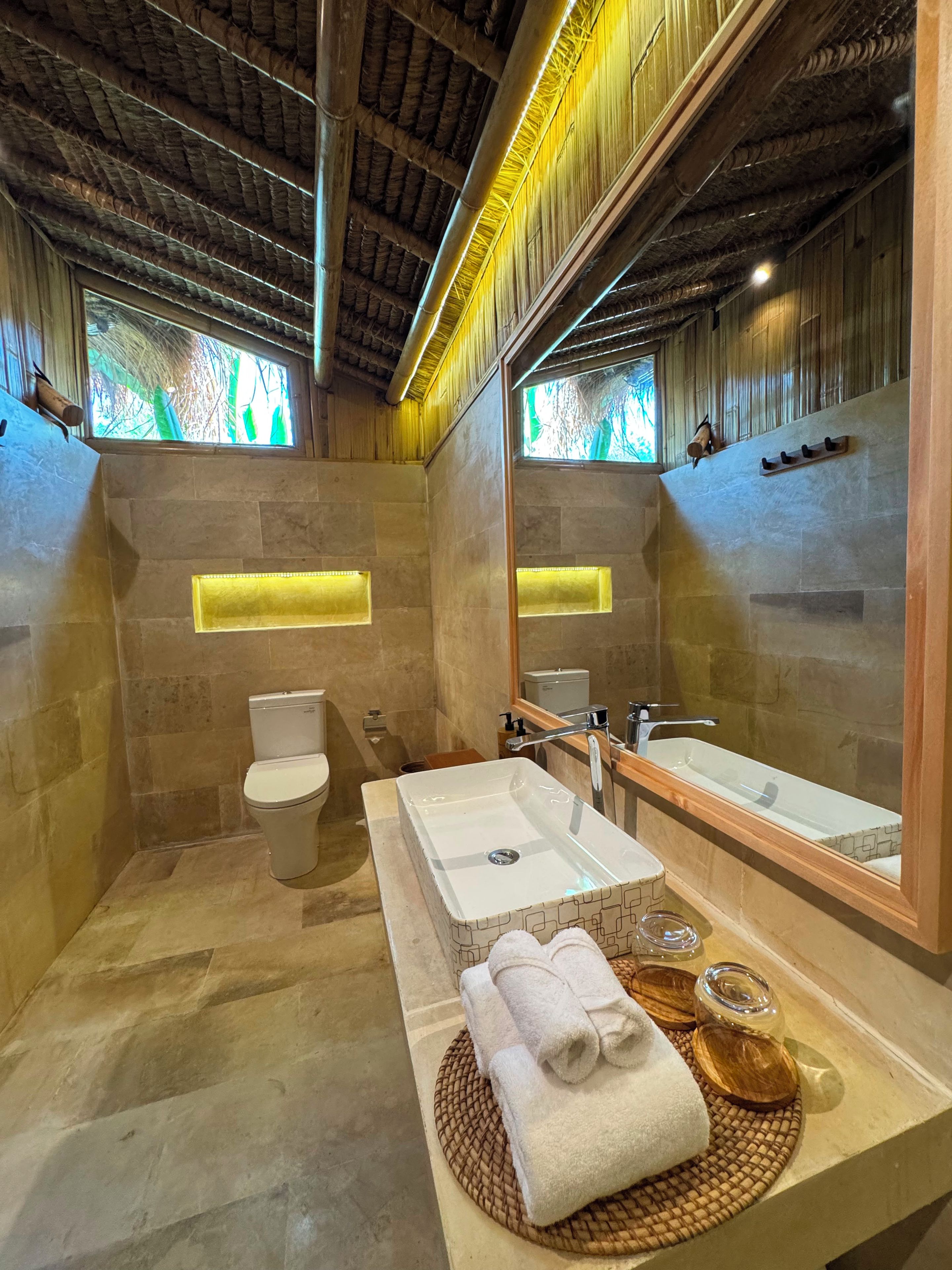 A modern bathroom with a stone wall, a white sink, a large mirror, and a wooden ceiling.