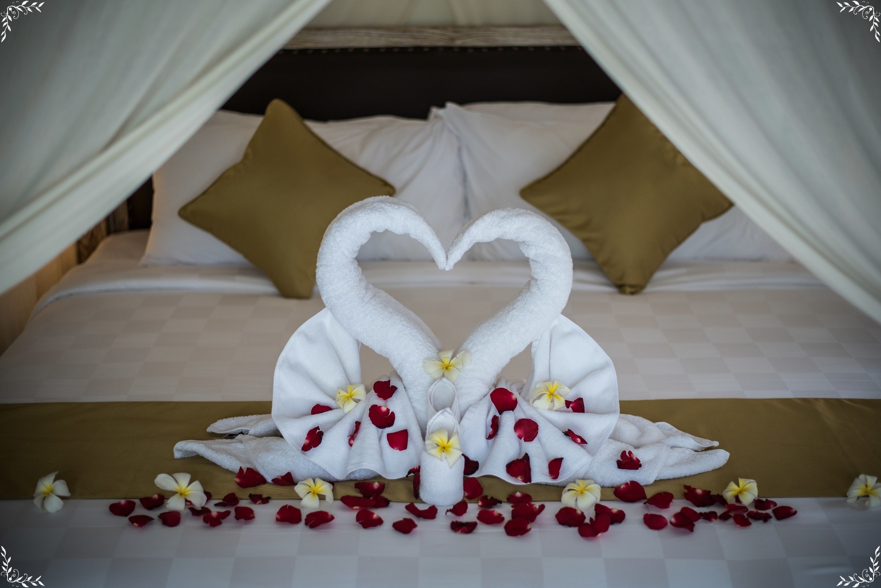 A bed with a canopy, white pillows, and a heart-shaped towel arrangement with flowers.