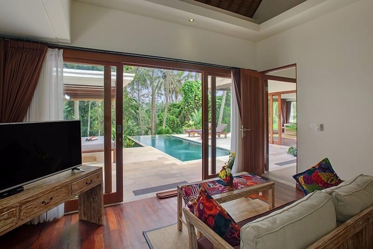 A living room with a television, a sofa, and a coffee table. There is a sliding glass door leading to a pool area.