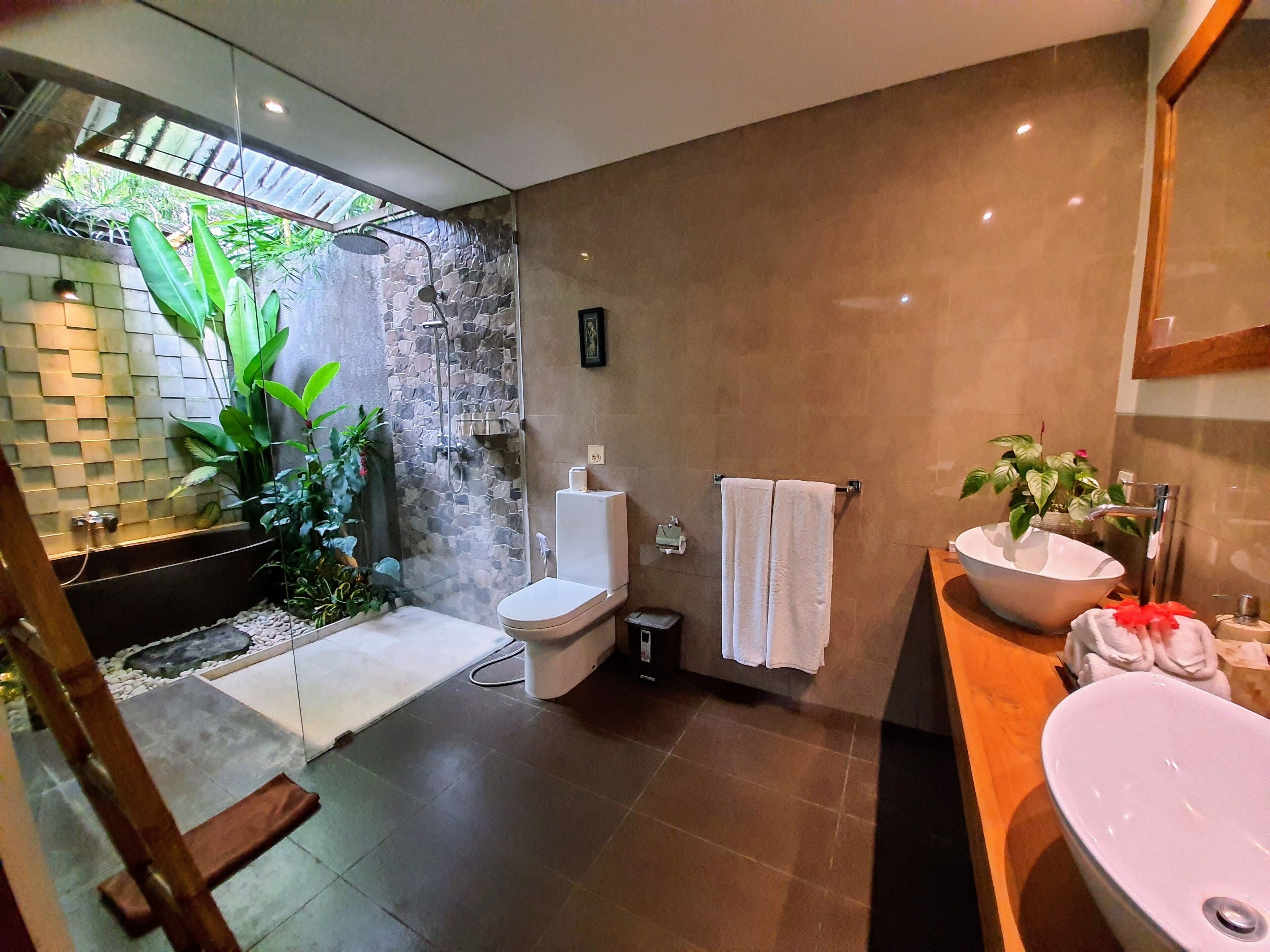 A modern bathroom with a stone shower wall, a white toilet, a large sink, and a small trash bin.