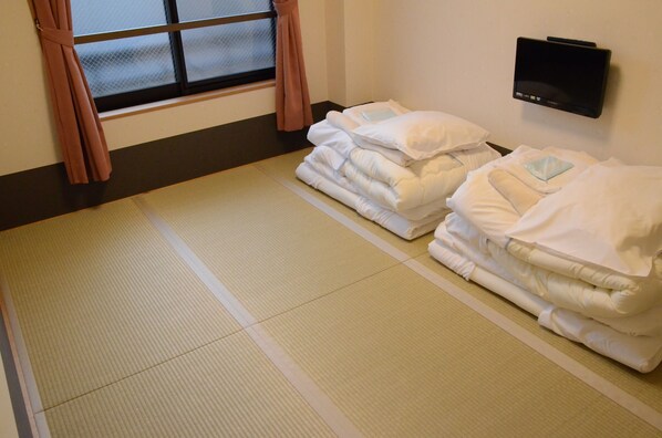 A room with tatami mats, a bed with white bedding, a TV on the wall, and a window with curtains.